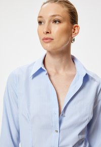 Camisa de botones de rayas azul claro y blanca con cuello, que presenta una textura suave y botones de metal, llevada con un pendiente sencillo.