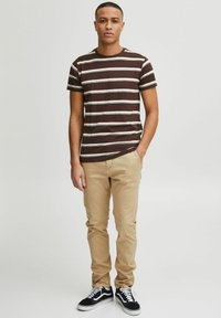 Brun och vit randig t-shirt, i bomullsblandning, med rund halsringning. Kombination med beige chinos och svarta sneakers, stående mot en enkel bakgrund.