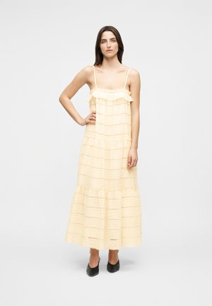 Ženska s ravnimi rjavimi lasmi pozira v brezrokavni večplastni maxi obleki svetlo rumene barve in črnih koničastih čevljih na belem ozadju.