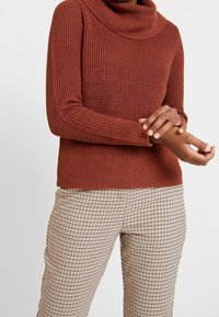Personne portant un pull à col roulé tricoté couleur rouille et un pantalon beige à carreaux, ajustant la manche de son bras gauche devant un fond uni.
