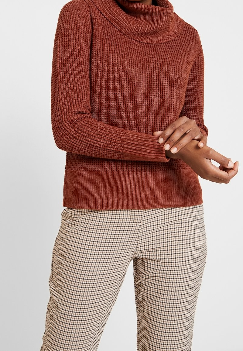 Personne portant un pull à col roulé tricoté couleur rouille et un pantalon beige à carreaux, ajustant la manche de son bras gauche devant un fond uni.