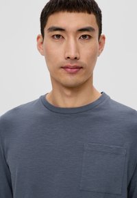 Szara koszula z długim rękawem, z okrągłym dekoltem i kieszonką na piersi. Materiał ma gładką teksturę, a kolor jest jednolity, bez wzorów.