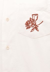 Λευκή τσέπη πουκαμίσου με καφέ κέντημα ενός τριαντάφυλλου που μπλέκεται με το μίσχο και τα φύλλα ενός ποτηριού κρασιού.