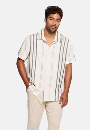 Hombre con cabello rizado que lleva una camisa de botones de manga corta en beige y blanco con rayas verticales, de pie con una mano en el bolsillo.