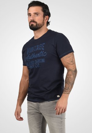 Navyblaue Baumwoll-T-Shirt mit einem Vintage-Druck in Blau. Kurze Ärmel, Rundhalsausschnitt und kontrastierende Steppnähte am Saum.