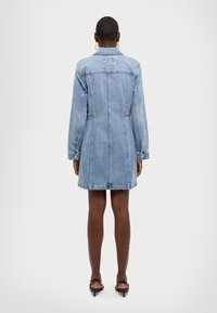 Lyseblå denimkjole med lange ermer, krage og sidelommer. Har synlige sømmer og gullknappeakenter ved ermkantene.