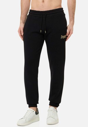 Mann trägt schwarze Jogginghose mit Kordelzugbund und weiße Sneakers, steht vor einfarbigem Hintergrund.