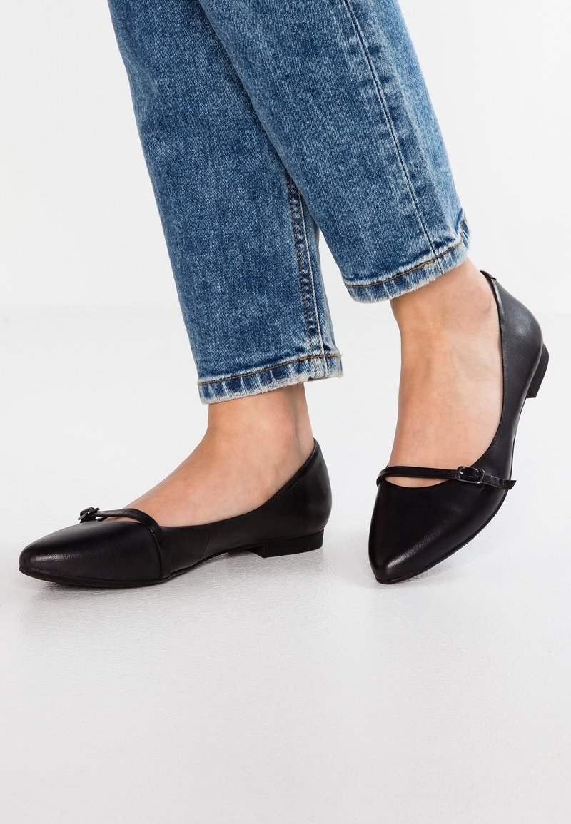 Personne portant un jean bleu et des chaussures plates noires à bout pointu avec une fine bride sur le dessus, debout sur un sol blanc lisse.