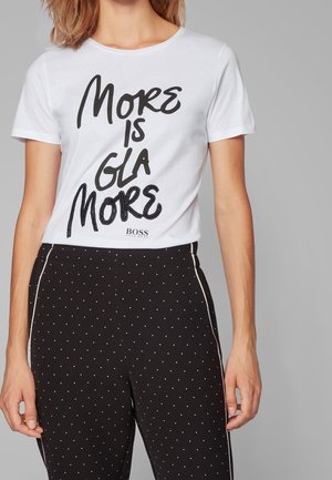 Mujer que lleva una camiseta blanca con el texto "More is Glamore" y pantalones negros con pequeños lunares blancos y rayas blancas laterales.