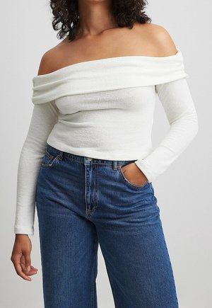 Osoba ubrana w białą bluzkę z dekoltem odsłaniającym ramiona i długim rękawem oraz dżinsy z wysokim stanem, z jedną ręką w kieszeni, na jednolitym tle.