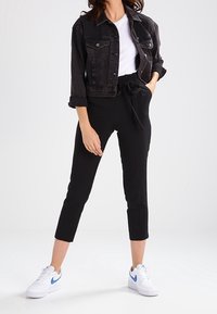 Svart denimjacka med upprullade ärmar, vit t-shirt, svarta tapered byxor och vita sneakers med blå detaljer. Ren och avslappnad outfitdesign.