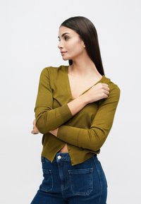 Cardigan vert moutarde à manches longues, col en V, boutonnage sur le devant, et poignets côtelés. Porté avec un jean bleu taille haute, avec poches arrière.