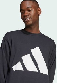 Sort sweatshirt med rund hals, der har et stort hvidt Adidas-logo-grafik på forsiden. Stoffet ser blødt ud med en afslappet pasform.