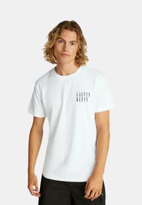 Camiseta blanca de algodón con mangas cortas y cuello redondo, con el logo de "Calvin Klein" impreso en el lado izquierdo del pecho en negro.