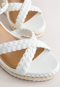 Sandale blanche avec brides tressées et semelle plateforme enveloppée de jute sur un fond clair.