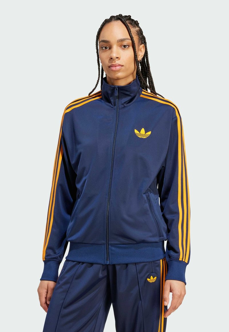 Marineblaues Zip-Jackett mit orangefarbenen Streifen entlang der Ärmel und Seiten. Mit einem kleinen gelben Markenlogo auf der Brust.