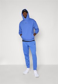 Blue Nike hoodie with a front pocket, matching blue joggers, and white sneakers. Features a black logo accent on the hoodie and pants.