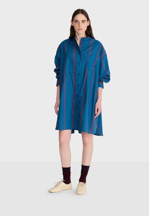 Vrouw staand in een losse blauwe overhemdjurk met verticale rode strepen, donkere sokken en crèmekleurige veterschoenen op een effen achtergrond.