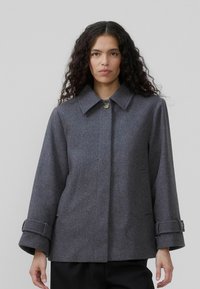 En kvinde med langt, krøllet hår bærer en grå, oversized frakke med krave og knap. Baggrunden er neutral og minimal.
