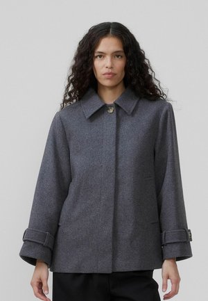 Una donna con lunghi capelli ricci indossa un cappotto grigio oversize con colletto e bottone. Lo sfondo è neutro e minimale.