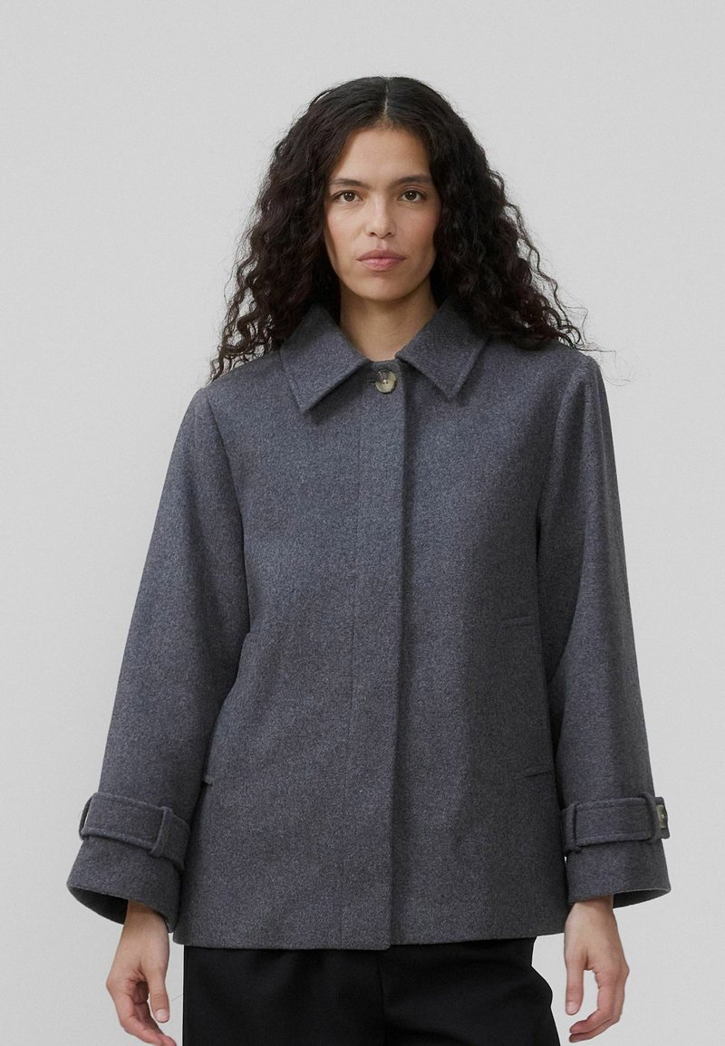 En kvinde med langt, krøllet hår bærer en grå, oversized frakke med krave og knap. Baggrunden er neutral og minimal.