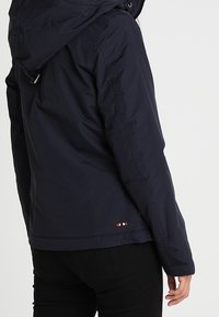 Veste bleu marine avec capuche, en matériau lisse et résistant à l'eau, dotée d'un petit logo avec des accents rouge, blanc et bleu près de l'ourlet.