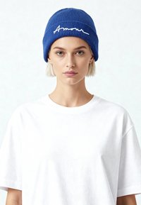 Cuffia a maglia blu con texture a coste e testo "Amour" ricamato in bianco, abbinata a una T-shirt bianca oversize semplice.