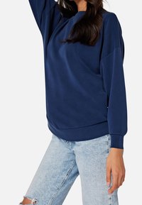 Felpa blu navy con vestibilità rilassata, collo rotondo e maniche lunghe; abbinata a jeans chiari strappati. Texture del tessuto liscia.