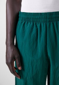 Calças leves em tom teal, com um cós elástico, apresentando um acabamento texturizado e detalhe franzido. Close-up de uma mão descansando sobre o tecido.