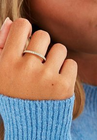 Goldfarbiger Ring mit eingelegten kleinen klaren Steinen, der an einer Hand getragen wird. Im Hintergrund ist ein blauer Rippensweater zu sehen.