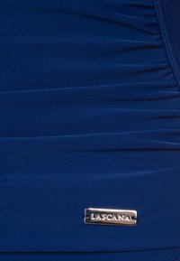 Tissu bleu marine avec texture froncée, arborant une étiquette en métal argenté portant la mention "LASCANÄ" de manière bien visible.