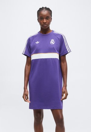 Jeune femme portant une robe violette Real Madrid Adidas avec des bandes blanches et des logos, debout devant un fond clair uni.
