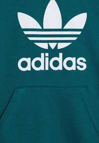 Teal sweatshirt featuring a white Adidas logo and trefoil graphic. It has a front pocket and a soft, textured fabric.