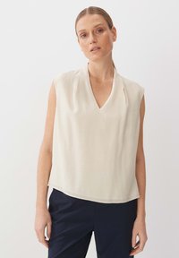 Blusa senza maniche color crema con scollo a V e sottili pieghe sulle spalle, caratterizzata da una texture liscia e una vestibilità rilassata e ampia.