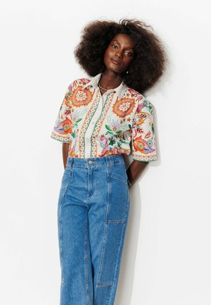Femme aux cheveux bouclés et volumineux, portant un chemisier à manches courtes coloré à motif floral et un jean bleu taille haute, debout devant un fond blanc.