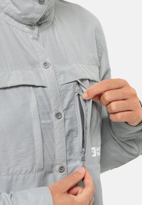 Hellgraue Jacke mit Knöpfen auf der Vorderseite, zwei Brusttaschen und einem Reißverschlussfach. Der Stoff sieht glatt und leicht aus.