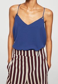 Haut sans manches bleu avec un décolleté en V et des bretelles réglables. Le mannequin porte un pantalon rayé beige et bordeaux avec des poches.