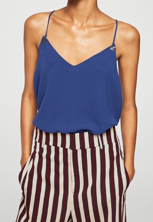 Haut sans manches bleu avec un décolleté en V et des bretelles réglables. Le mannequin porte un pantalon rayé beige et bordeaux avec des poches.