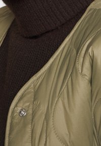 Veste matelassée en duvet vert olive avec un design structuré et des boutons-pression argentés, portée sur un pull col roulé côtelé marron foncé.