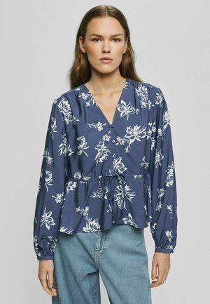 Woman wearing a blue long-sleeve blouse with white floral print and drawstring waist paired with light blue jeans, standing against a plain background.