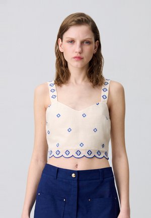 Top corto beige con ricami blu a forma di diamante e motivi floreali. Presenta spalline larghe e un orlo ondulato. Il materiale sembra leggero.