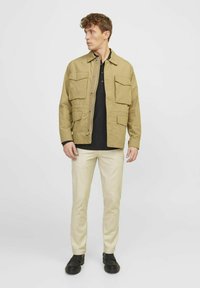 Beige Utility-Jacke mit knöpfbaren Taschen, getragen über einem schwarzen Poloshirt, kombiniert mit beigen Hosen und schwarzen Schuhen, vor einem weißen Hintergrund stehend.