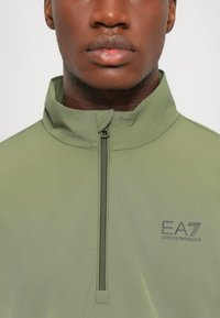 Lys olivengrønn quarter-zip genser med høy krage; har en glatt tekstur og EA7 Emporio Armani-logoen trykt på brystet.