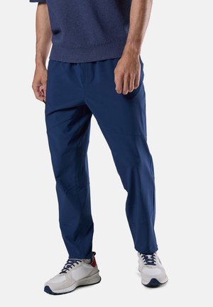 Mann trägt marineblaue Hose, weiße Sneaker mit marineblauen und roten Details und ein dunkelblaues, kurzärmeliges Strickoberteil, steht vor einem einfarbigen Hintergrund.