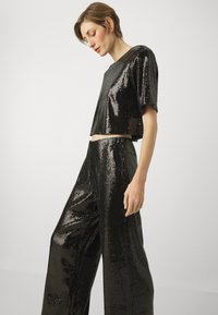Fekete flitteres crop top és bő szárú nadrág, fényes textúrával és rövid ujjakkal. Az anyag megcsillan a fényben, ragyogó hatásokat keltve.