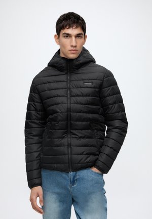 Jeune homme portant une veste matelassée noire à capuche avec fermeture éclair et un jean bleu, debout devant un fond clair uni.