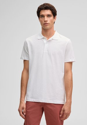 Junger Mann mit kurzen lockigen Haaren, der ein einfaches weißes kurzärmeliges Poloshirt und rostfarbene Hosen trägt, vor einem grauen Hintergrund stehend.