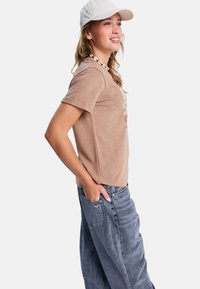Jonge vrouw die glimlacht, gekleed in een beige t-shirt, blauwe jeans, een witte schelpenhalsketting en een lichtgekleurde baseballcap, staand in een relaxte houding.