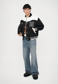 Giacca varsity bianca e nera con una lettera "M" ricamata, abbinata a jeans svasati blu e scarpe nere ricamate, con una piccola borsa in mano.