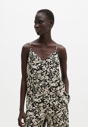 Jeune femme aux cheveux courts portant un haut sans manches à fleurs noir et blanc et un pantalon large assorti sur un fond blanc uni.